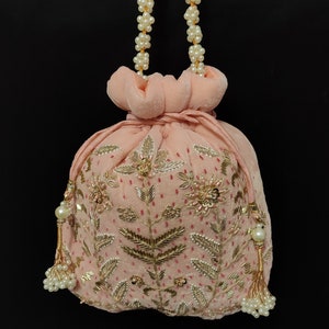 Puede incluir: Una bolsa de cord&oacute;n de terciopelo rosa con bordados dorados y plateados y detalles de perlas. La bolsa tiene una correa de cadena de perlas.