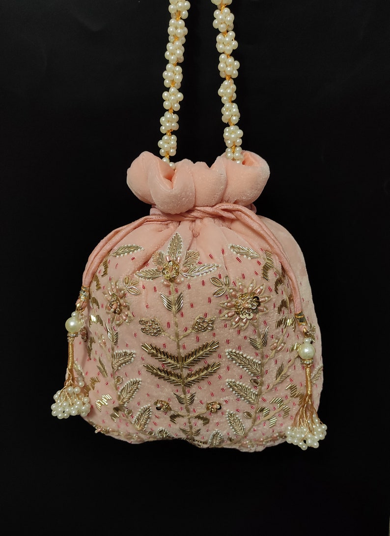 Puede incluir: Una bolsa de cord&oacute;n de terciopelo rosa con bordados dorados y plateados y detalles de perlas. La bolsa tiene una correa de cadena de perlas.