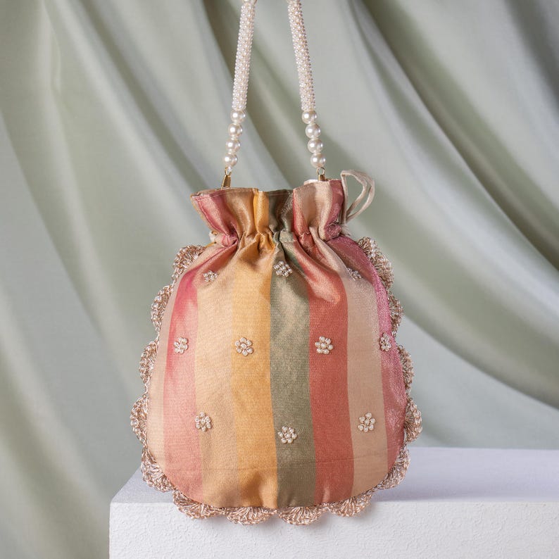 May include: A colorful, striped drawstring bag with a beaded handle. The bag features vertical stripes in shades of pink, yellow, and green, adorned with small, flower-shaped embellishments. The edges are trimmed with a delicate, crocheted border.