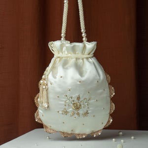 Puede incluir: Bolso de satén marfil con cordón, asas y borla de perlas. El bolso presenta bordados dorados, lentejuelas y un borde festoneado. Las perlas dispersas realzan el diseño elegante. Un accesorio ideal para ocasiones especiales.