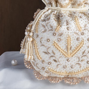 May include: An ivory drawstring bag, adorned with gold and pearl beadwork. The bag showcases floral and leaf designs, a scalloped edge, and a pearl tassel. A single pearl sits on the white surface beside the bag.