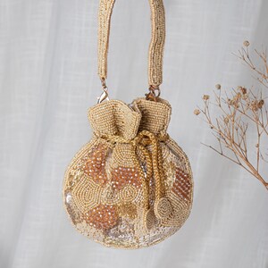 May include: A beige beaded drawstring bag with a gold chain and handles. The bag features a pattern of gold and beige beads and sequins. The handles and drawstring are also beaded. The bag is hanging against a white background.