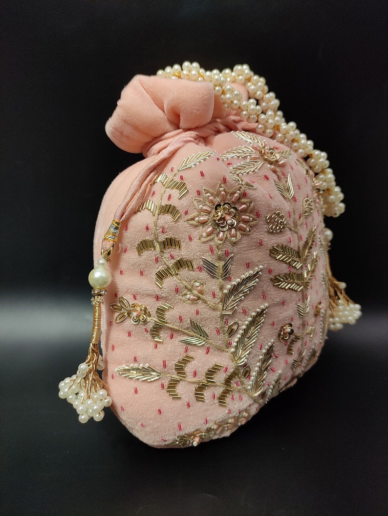 Puede incluir: Una bolsa de cord&oacute;n de terciopelo rosa con adornos dorados y de perlas. La bolsa est&aacute; decorada con bordados florales y tiene un borla de perlas.