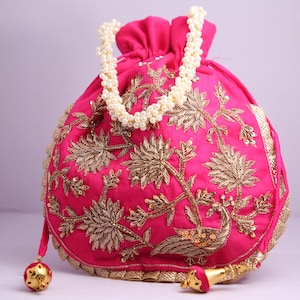 Puede incluir: Una bolsa de cordón rosa con bordados dorados y un asa de collar de perlas. La bolsa presenta un diseño floral con un pavo real en el centro.