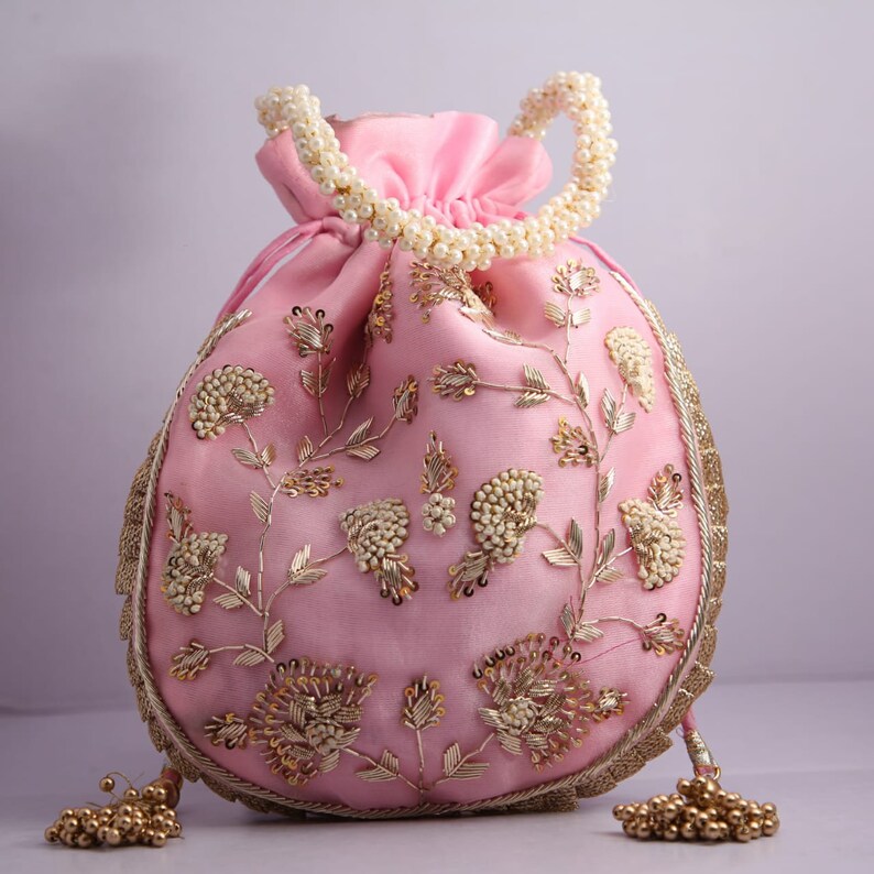 May include: A pink drawstring bag with gold and white embroidery. The bag has a pearl handle and gold tassel accents.