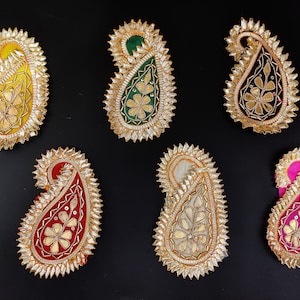 Könnte beinhalten: Sechs paisleyförmige Stoffpatches mit goldenen und silbernen Verzierungen. Die Patches sind in verschiedenen Farben, darunter Gelb, Grün, Schwarz, Rot, Weiß und Pink.