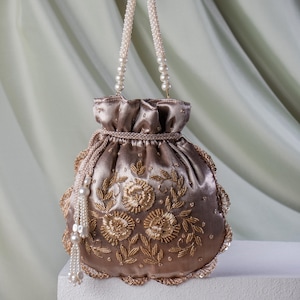 May include: A taupe satin drawstring bag with gold floral embroidery and a beaded handle. The bag features a scalloped edge and a tassel of pearls. The bag is approximately 18 cm tall.