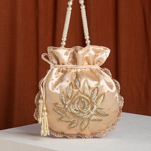 May include: A champagne-colored satin drawstring bag with a floral embroidery design in gold and silver thread. The bag features a beaded handle and a tassel with pearl accents. The edges are trimmed with a delicate lace.