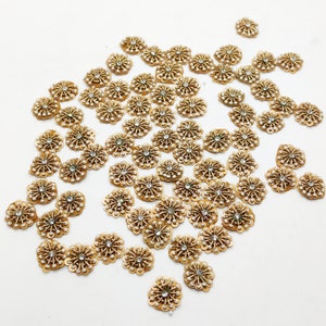 Può includere: Un grande gruppo di decorazioni dorate a forma di fiore con accenti di perle bianche. Le decorazioni sono sparse su una superficie bianca.