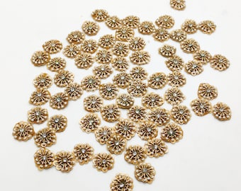 May include: A large group of gold-colored flower-shaped embellishments with white pearl-like accents. The embellishments are scattered on a white surface.
