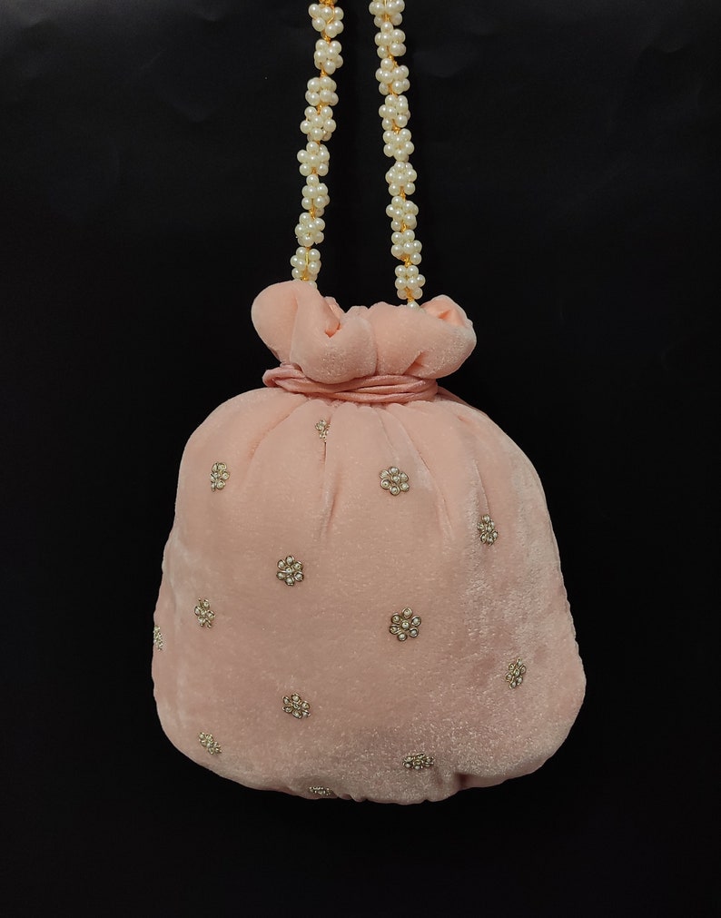 Puede incluir: Una bolsa de cord&oacute;n de terciopelo rosa con una correa de cadena de perlas y adornos dorados. La bolsa est&aacute; decorada con peque&ntilde;os acentos circulares dorados.