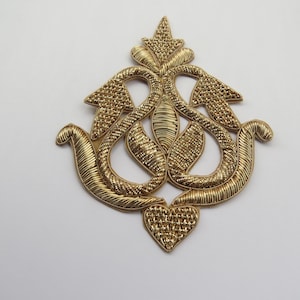 Può includere: Un designo decorativo intricato di colore dorato con una forma di cuore nella parte inferiore. Il designo è fatto di fili intrecciati e ha uno stile tradizionale e ornato.