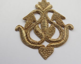 Op de afbeelding: Een ingewikkeld, goudkleurig decoratief ontwerp met een hartvorm aan de onderkant. Het ontwerp is gemaakt van geweven draden en heeft een traditionele, sierlijke stijl.