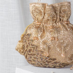 Könnte beinhalten: Eine goldfarbene Beuteltasche mit aufwendiger Perlenarbeit und gestickten Blumenmustern. Die Tasche hat fächerförmige Verzierungen und baumelnde Perlenakzente. Die Tasche ist aus schimmerndem Stoff gefertigt und hat eine Quaste an der Seite.