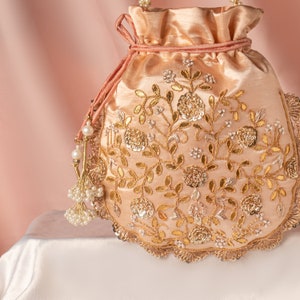 Könnte beinhalten: Eine pfirsichfarbene Kordeltasche mit goldenen und silbernen Stickereien und Perlenakzenten. Die Tasche hat einen gewellten Rand und einen Quasten.