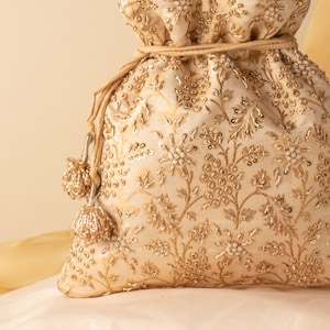 May include: A cream-coloured drawstring bag with gold embroidery and beaded tassels. The bag is decorated with a floral pattern and has a delicate, feminine feel.