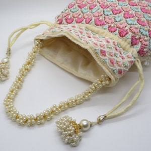 以下が含まれることがあります： ピンクと水色の刺繍が施されたアイボリー色の巾着袋。白いビーズで装飾され、パールビーズの紐とタッセルが付いています。エレガントで装飾的なアクセサリーです。