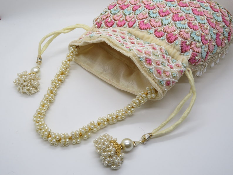 以下が含まれることがあります： ピンクと水色の刺繍が施されたアイボリー色の巾着袋。白いビーズで装飾され、パールビーズの紐とタッセルが付いています。エレガントで装飾的なアクセサリーです。