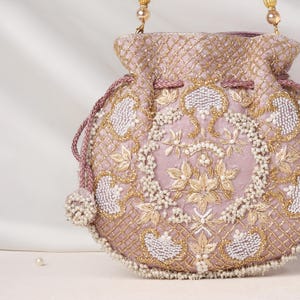 Può includere: Una borsa a sacchetto color malva con chiusura a coulisse, decorata con ricami in filo dorato e perline bianche in un motivo a reticolo. Il davanti presenta un motivo floreale centrale circondato da bordi decorativi e nappe di perline.