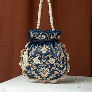 May include: A navy blue drawstring bag with intricate gold floral embroidery and sequin detailing. The bag features pearl beaded handles and a tassel, offering an elegant and ornate design. The bag is sitting on a white surface.