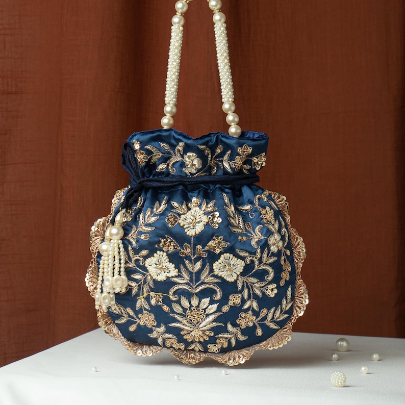 May include: A navy blue drawstring bag with intricate gold floral embroidery and sequin detailing. The bag features pearl beaded handles and a tassel, offering an elegant and ornate design. The bag is sitting on a white surface.