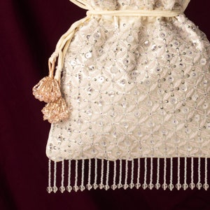 May include: A white drawstring bag with a beaded design and a fringe trim. The bag has a tassel on the side.