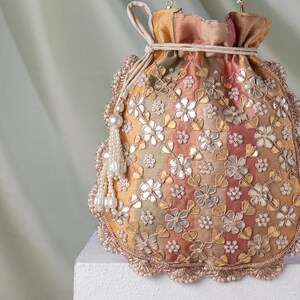 May include: A decorative drawstring pouch in shades of gold, peach, and green, adorned with floral embroidery and mirror accents. The pouch features a beaded tassel and a scalloped edge, creating an elegant accessory.