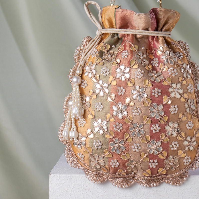 May include: A decorative drawstring pouch in shades of gold, peach, and green, adorned with floral embroidery and mirror accents. The pouch features a beaded tassel and a scalloped edge, creating an elegant accessory.