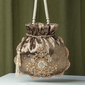 Może przedstawiać: Złota torebka ze sznurkiem, z uchwytem z koralików i misternym haftem kwiatowym. Torba ma falisty brzeg i jest ozdobiona małymi, błyszczącymi zdobieniami. Z boku wisi koralikowa frędzel, dodając do jej ozdobnego designu.