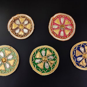 May include: Five round embroidered patches with floral designs. The patches are in various colors, including green, blue, pink, and orange. Each patch has a gold center with a small, round, clear gem.