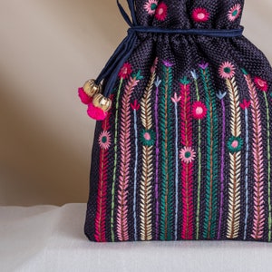 May include: A dark blue drawstring bag with a floral embroidery design. The bag has a tassel with gold beads and pink pom-poms.