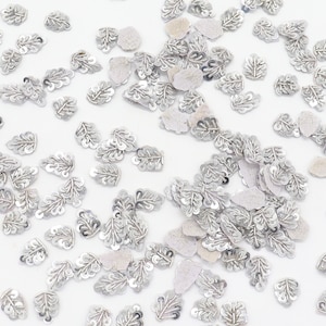 May include: A scattering of silver leaf-shaped sequins on a white background. The sequins are made of a shiny material and have a delicate, intricate design.