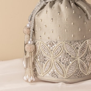 May include: A cream-colored drawstring bag with a beaded design. The bag has a silver and white beaded trim and a tassel with white beads.