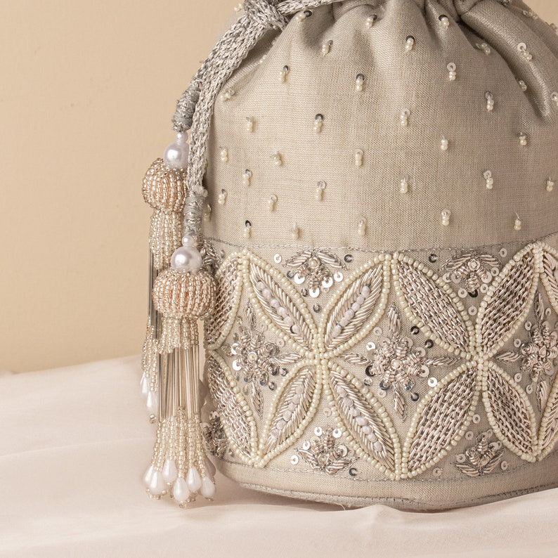 May include: A cream-colored drawstring bag with a beaded design. The bag has a silver and white beaded trim and a tassel with white beads.