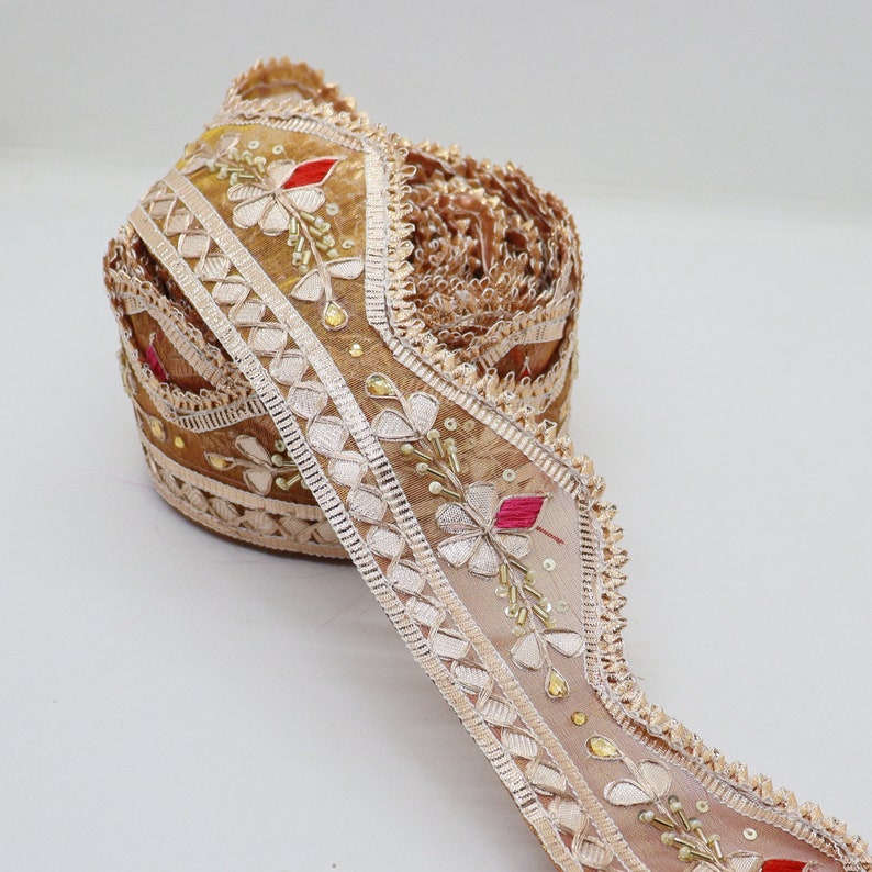 May include: A roll of beige embroidered trim with a scalloped edge. The trim features floral designs with red, pink, and green accents. The trim is made of a sheer fabric with a delicate texture.