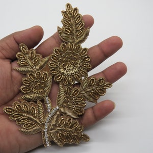 以下が含まれることがあります： ビーズとスパンコールで飾られた花柄の金色の刺繍アップリケ。アップリケには、3つの花と葉が付いた枝があります。
