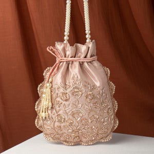 May include: A blush-colored drawstring bag with intricate gold floral embroidery. The bag features a scalloped edge, a pearl-beaded handle, and a tassel. The bag is made of a silky material and is suitable for special occasions.