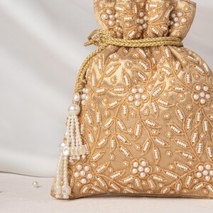 Puede incluir: Una bolsa dorada con cierre de cordón que presenta un intrincado bordado con cuentas blancas y lentejuelas dispuestas en patrones florales y de enredaderas. Incluye un cordón dorado y una borla hecha de cuentas blancas y perlas.