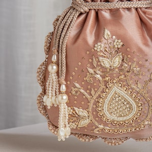 May include: A dusty rose-colored drawstring bag with intricate gold embroidery and pearl accents. The bag features a scalloped edge and decorative tassels with large pearl beads. The design includes floral and paisley-inspired patterns.