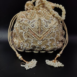 May include: A beige beaded drawstring bag with intricate floral and geometric patterns. The bag features white and pink accents, a beaded handle, and decorative tassels. The bag is embellished with sequins and pearls, creating a luxurious look.