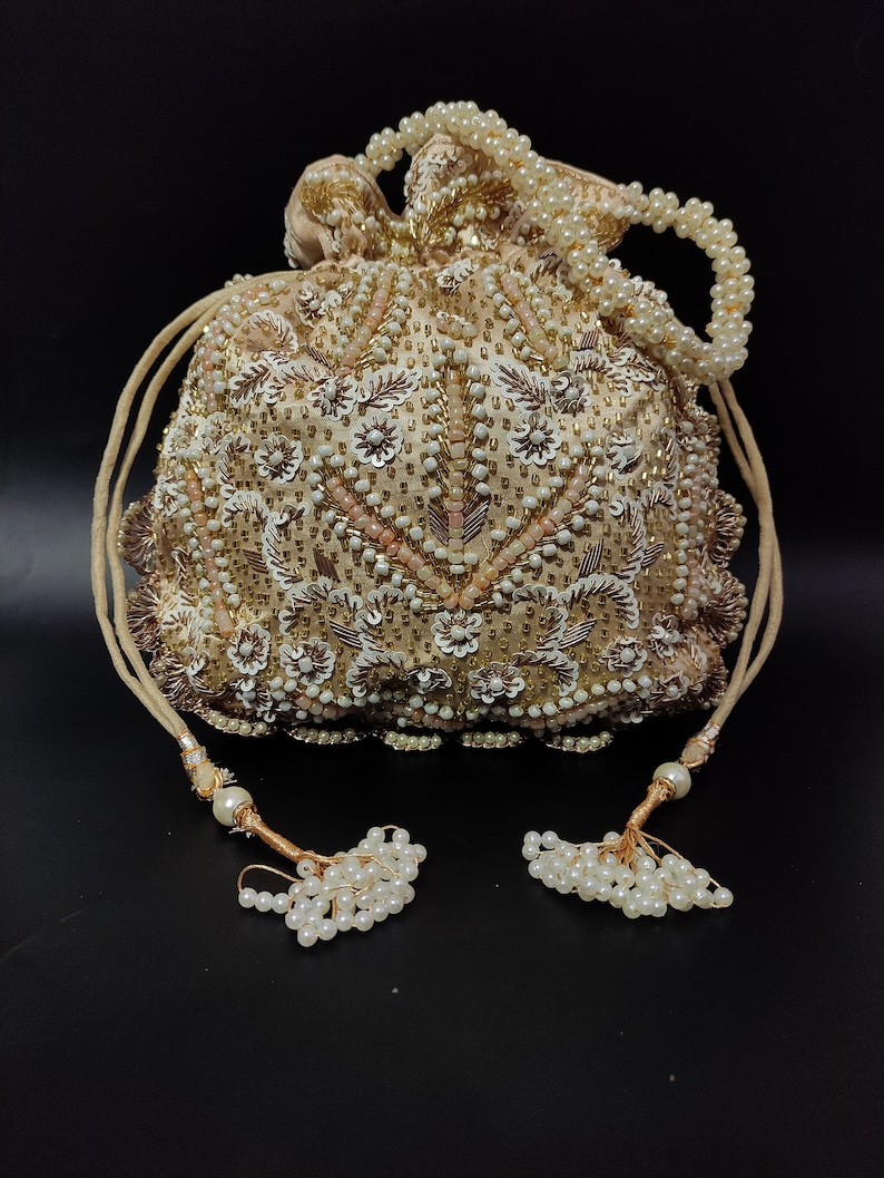 May include: A beige beaded drawstring bag with intricate floral and geometric patterns. The bag features white and pink accents, a beaded handle, and decorative tassels. The bag is embellished with sequins and pearls, creating a luxurious look.