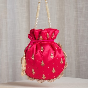 May include: A vibrant fuchsia satin pouch bag with a drawstring closure and pearl beaded handles. The bag is adorned with gold embroidery and a scalloped gold trim. A pearl tassel hangs from the side.