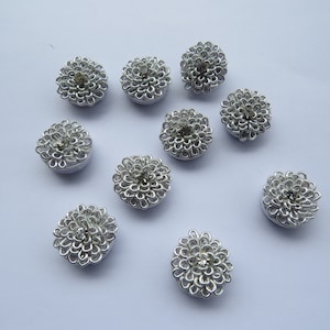 Puede incluir: Diez botones de metal de color plateado en forma de flor con centros de diamantes de imitación transparentes. Los botones están dispuestos en semicírculo sobre un fondo blanco.