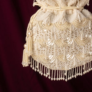 May include: A white beaded drawstring bag with a pearl tassel. The bag is decorated with a pattern of white and gold beads.