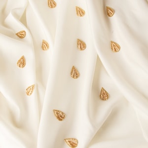 Könnte beinhalten: Weißer Stoff mit subtilem Glanz, verziert mit goldenen, blattförmigen Verzierungen. Der Stoff ist drapiert und gefaltet und erzeugt eine weiche, fließende Textur.