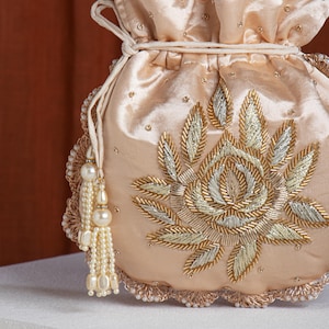 Peut inclure: Une pochette à cordon de serrage pêche avec une broderie florale dorée et argentée complexe. La pochette présente un gland perlé et un bord festonné avec de petites perles. Le design est symétrique et élégant, adapté aux occasions spéciales.