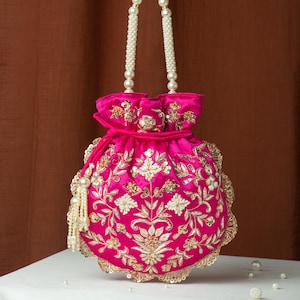 May include: A hot pink, round handbag with intricate gold embroidery and a pearl beaded handle. The bag features a drawstring closure and a scalloped edge, with a pearl tassel. The bag is approximately 8 inches in diameter.