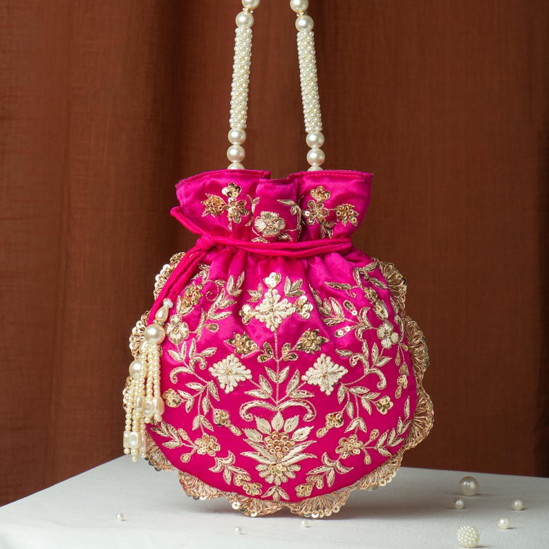 May include: A hot pink, round handbag with intricate gold embroidery and a pearl beaded handle. The bag features a drawstring closure and a scalloped edge, with a pearl tassel. The bag is approximately 8 inches in diameter.