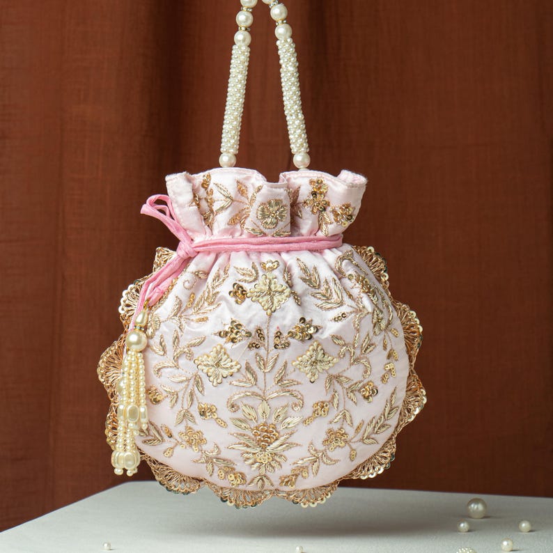 May include: A light pink, round drawstring bag with intricate gold embroidery and sequin details. The bag features a beaded handle and a pink drawstring closure. A gold tassel with pearls hangs from the side. The bag is likely for special occasions.