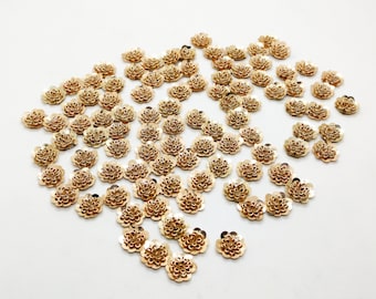 May include: A collection of small, gold-colored flower-shaped embellishments scattered on a white surface. Each flower has multiple petals and a detailed center. These decorative elements are suitable for various craft projects.